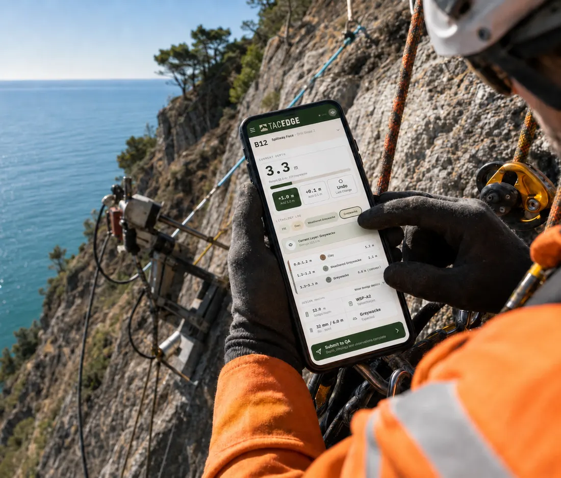 Operator on ropes at a coastal rock face recording a drill log on a phone in TACEDGE, drill rig visible behind